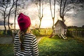 girl in a striped sweater and a red hat looks at a playground in the garden