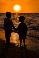 Child girl and boy holding hands in front of sea at sunset