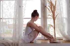 Ballerina in beautiful white dress and shoes, sitting near the white window