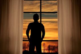 Silhouette of a man, looking on the colorful and beautiful sunset, behind the trees, through the window with the curtain