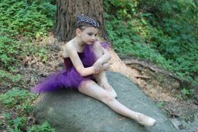 young ballerina in a purple dress sits on a stone in the forest