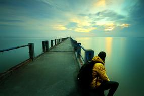 Thinking man, with the backpack, sitting on the beautiful pier, among the ocean, at colorful and beautiful sunset