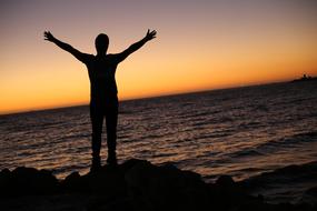 silhouette of a free man with raised hands on a rocky shore against a sunset background