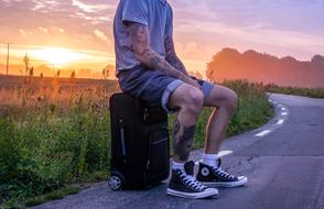 Traveler with the tattoos, sitting on the suitcase, on the road, among the colorful fields, at colorful and beautiful sunrise