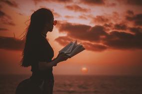 Girl in glasses, reading a book on the beach, at beautiful and colorful sunset