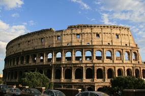 Beautiful Colosseum, among the cars and green trees, in Rome, Italy, at blue sky with clouds on background