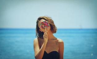 young girl holds colorful candy at face in front of Sea