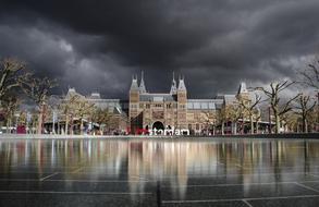 picturesque Rijksmuseum building mirroring on wet plaza beneath stormy clouds in Amsterdam