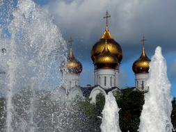 Beautiful fountain with water jets and church with golden domes, at blue sky with white clouds