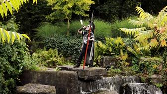 Roman soldier, with the weapon, on the rock with the waterfall, among the colorful plants