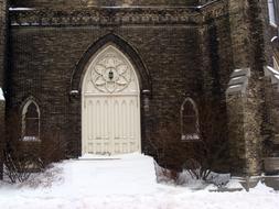 Door of the old church with windows, among the snow and plants, in the winter