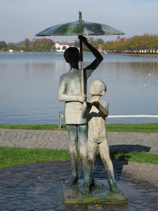 Statues of people with umbrella near the water, among the colorful plants, in Schwerin, Germany