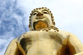 Low angle shot of the beautiful, shiny gold sculpture of Buddha, at blue sky with white clouds on background