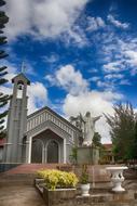 Beautiful landscape with the church and statue, among the colorful plants, under the blue sky with clouds