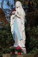 Beautiful Statue of Maria, with colorful flowers on the stand, near the colorful plants