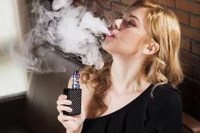 Beautiful, blonde girl, vaping electronic cigarette, on the sofa, near the brick wall