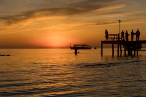 Silhouettes of the people, on the pier, above the beautiful water with the boat, at colorful and beautiful sunset,