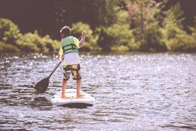 Boy in the green life vest, paddling on the beautiful lake, with ripple, in sunlight, near the colorful plants