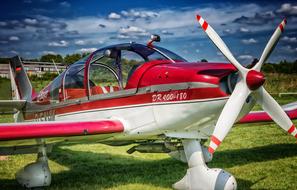 Red and white, shiny aircraft, with the propeller, on the green meadow, among the colorful plants, under the blue sky with clouds, in France