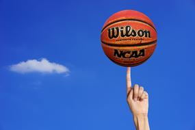 Beautiful, orange and black basketball, on the finger, at blue sky with white clouds on background