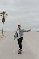 Bearded man in glasses, skateboarding on the path, among the sand, near the palm tree