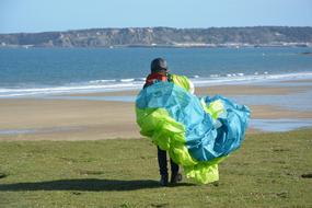 Paraglider, with the turquoise and lime parachute, on the green meadow, near the sandy beach, and mountains on background