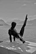 Black and white photo of the statue of the sculpture of the man, doing gymnastics, near the water, among the mountains