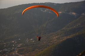 Person, doing paragliding on the orange and yellow, gradient parachute, above the beautiful and colorful mountains
