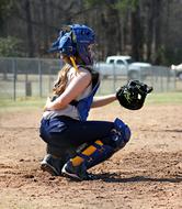 Softball girl catcher in equipment, on the sand field, with the fence, near the trees