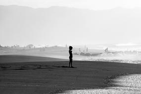 Black and white photo of the kid, on the beautiful, sandy beach and mountains in fog on background