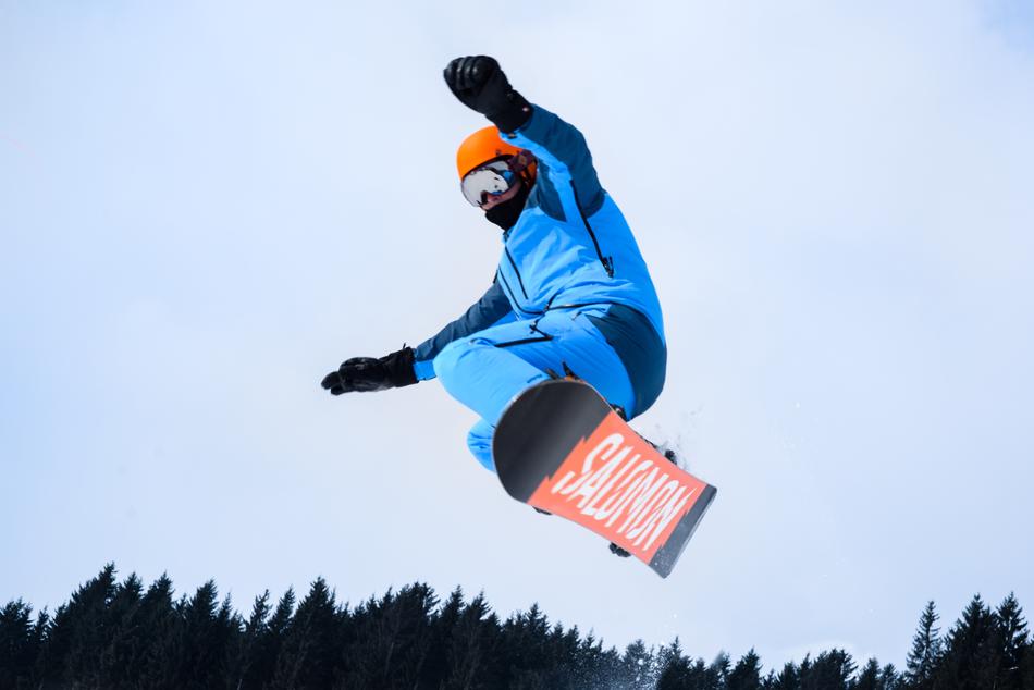 Colorful person in glasses, jumping on the snowboard in winter, near the trees, under the cloudy sky