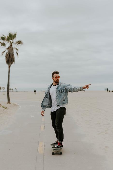 Bearded man in glasses, skateboarding on the path, among the sand, near the palm tree