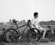 Black and white photo with the man, resting near the bike and fence, near the field