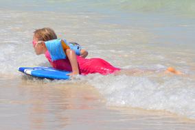 Blonde child girl, with the equipment, surfing on the sandy beach with water