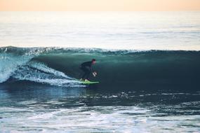 Surfer on the green surfboard, on the beautiful, green sea, with the wave, at colorful and beautiful sunset