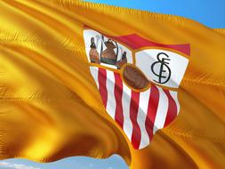 Colorful logo of Sevilla Fc of La Liga, on the gold flag, at blue sky with white clouds on background