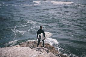 Surfer with surfboard, standing on the rocky shore with waves