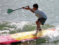 Boy surfing on the colorful surfboard, with paddle, on the water with waves in sunlight