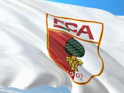 Colorful, wavy flag of FC Augsburg, in sunlight, at background with blue sky with clouds