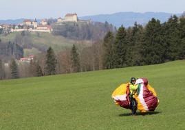 Paraglider with colorful parachute, on the beautiful, green meadow among the trees, near the buildings, in Switzerland