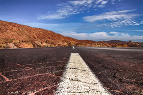Close-up of the road, with the white lines, among the colorful and beautiful hills, under the blue sky with white clouds