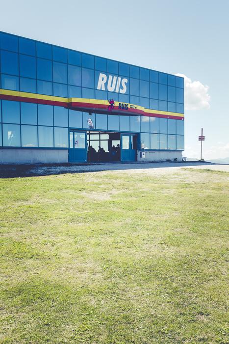 Shiny building with 'Ruis' sign near the green grass, under the blue sky with clouds, in the North Italy