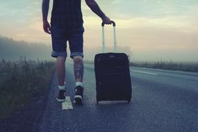 Traveler, concept, man with wheeled bag walking on road at dusk