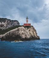 Lighthouse, on the beautiful, rocky shore, with the green plants, near the blue water, under the sky with clouds