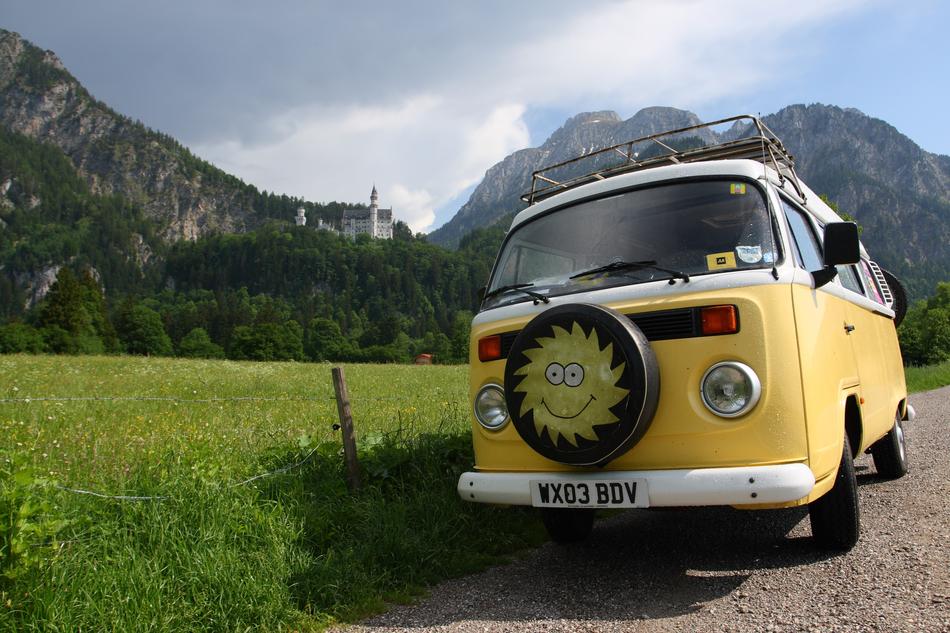 Beautiful, retro, yellow and white campersvan with the Sun near the green mountains and Neuschwanstein in Bavaria, Germany
