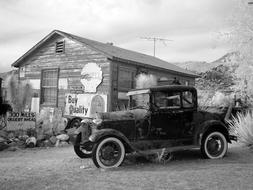 Black and white photo with the beautiful, vintage car near the building, among the plants, near the mountains