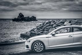 Black and white photo with shiny car, near the sea beach with rocks, under the clouds