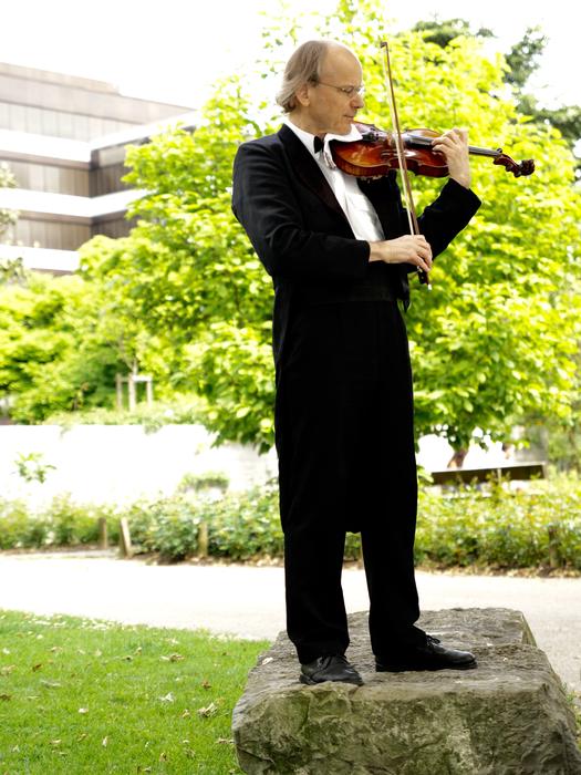 Man in suit, playing violin on the rock, among the colorful plants, near the building