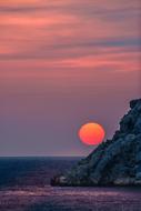 photo of a yellow-pink sun behind the rocks against the background of the evening sky in Greece