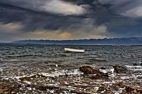 White boat on on the beautiful water with waves, view from the rocky coast, under the cloudy sky